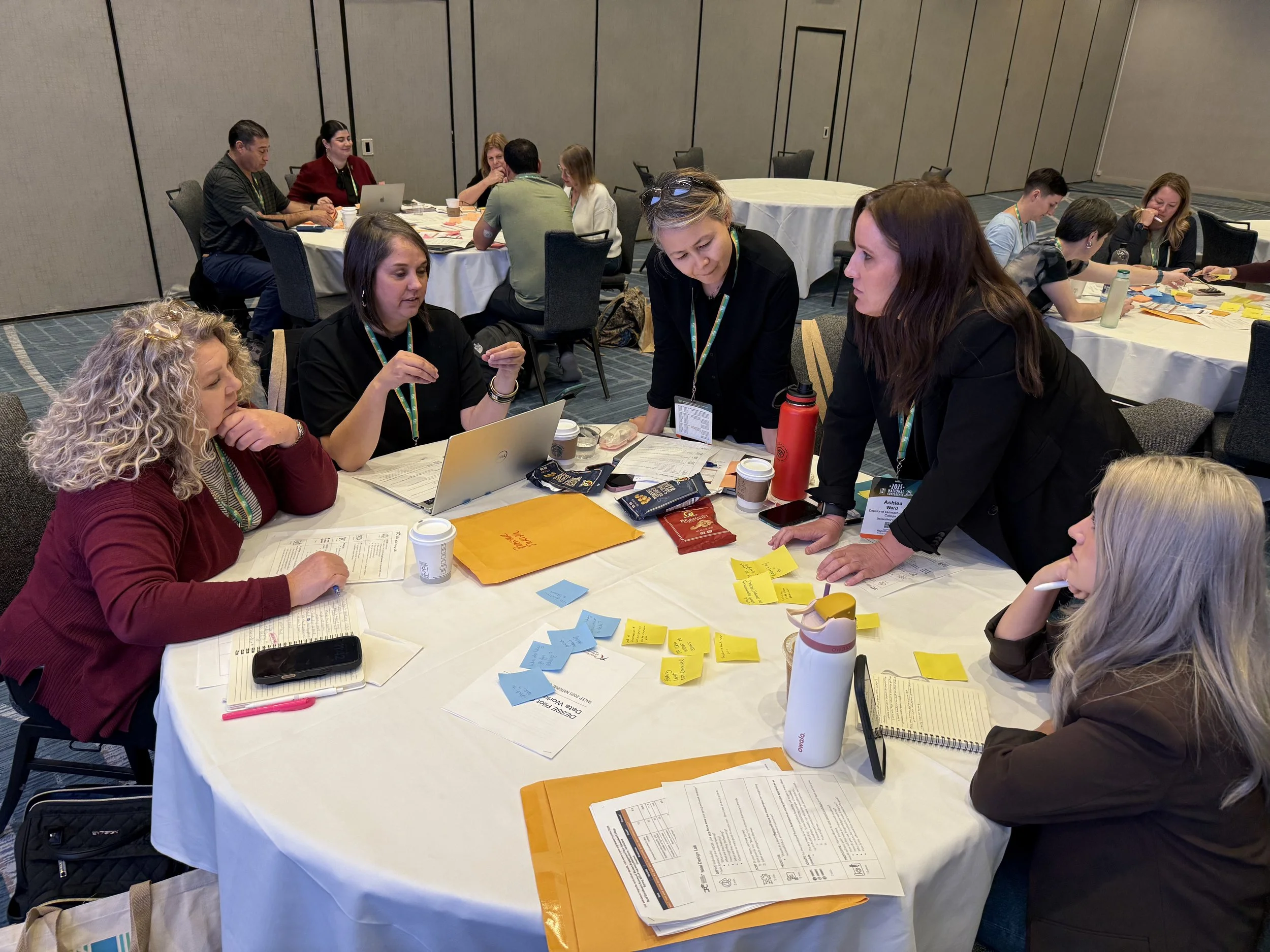 Group of women engaged in a discussion around a conference table with sticky notes, laptops, and papers.