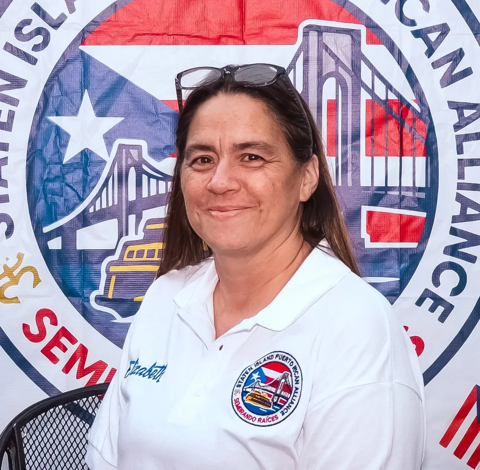 A woman in a white polo shirt with an embroidered badge on it, standing in front of a large banner with the SIPRA logo.