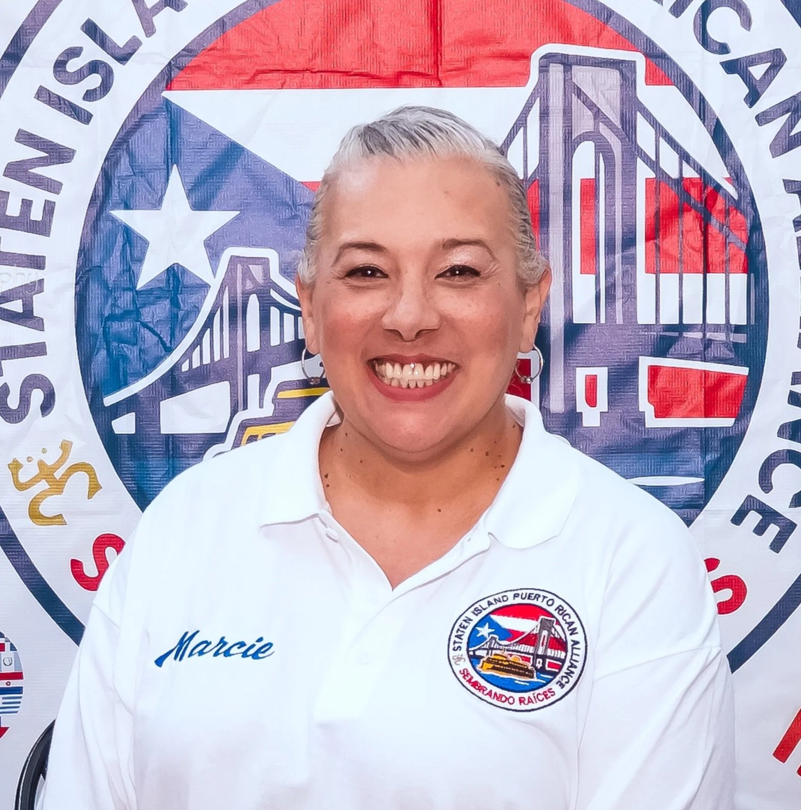 A woman smiling in front of a Puerto Rican flag and a seal reading 'Staten Island Puerto Rican Alliance.' She is wearing a white shirt with the name Marcie embroidered on it.