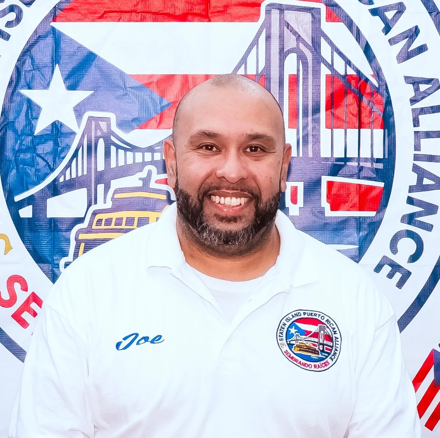 A smiling man with a beard wearing a white shirt with 'Joe' embroidered on it and a patch with a logo from the Staten Island Puerto Rican Alliance Logo, standing in front of a colorful mural featuring the Goethals Bridge and a Puerto Rican flag.
