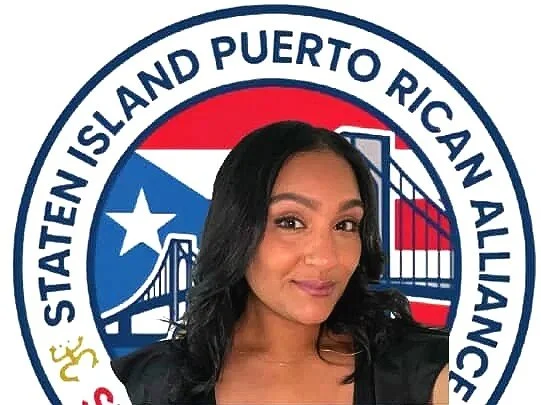 A woman with long black hair taking a selfie in front of a Puerto Rican American flag and a circular logo with the text "Statein Island Puerto Rican Alliance".