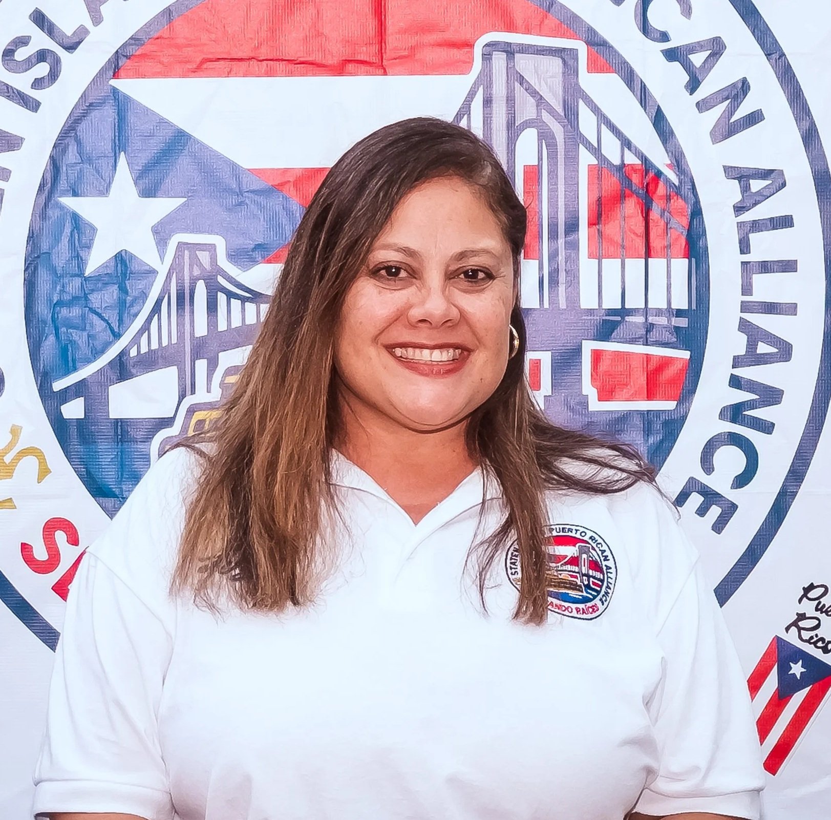 A woman with long brown hair smiling, wearing a white shirt with an embroidered logo, standing in front of a backdrop featuring the logo of the SIPRA, with an illustration of the Goethals Bridge and elements of the Puerto Rican flag.