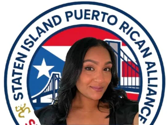 A woman with long black hair taking a selfie in front of a Puerto Rican American flag and a circular logo with the text "Statein Island Puerto Rican Alliance".