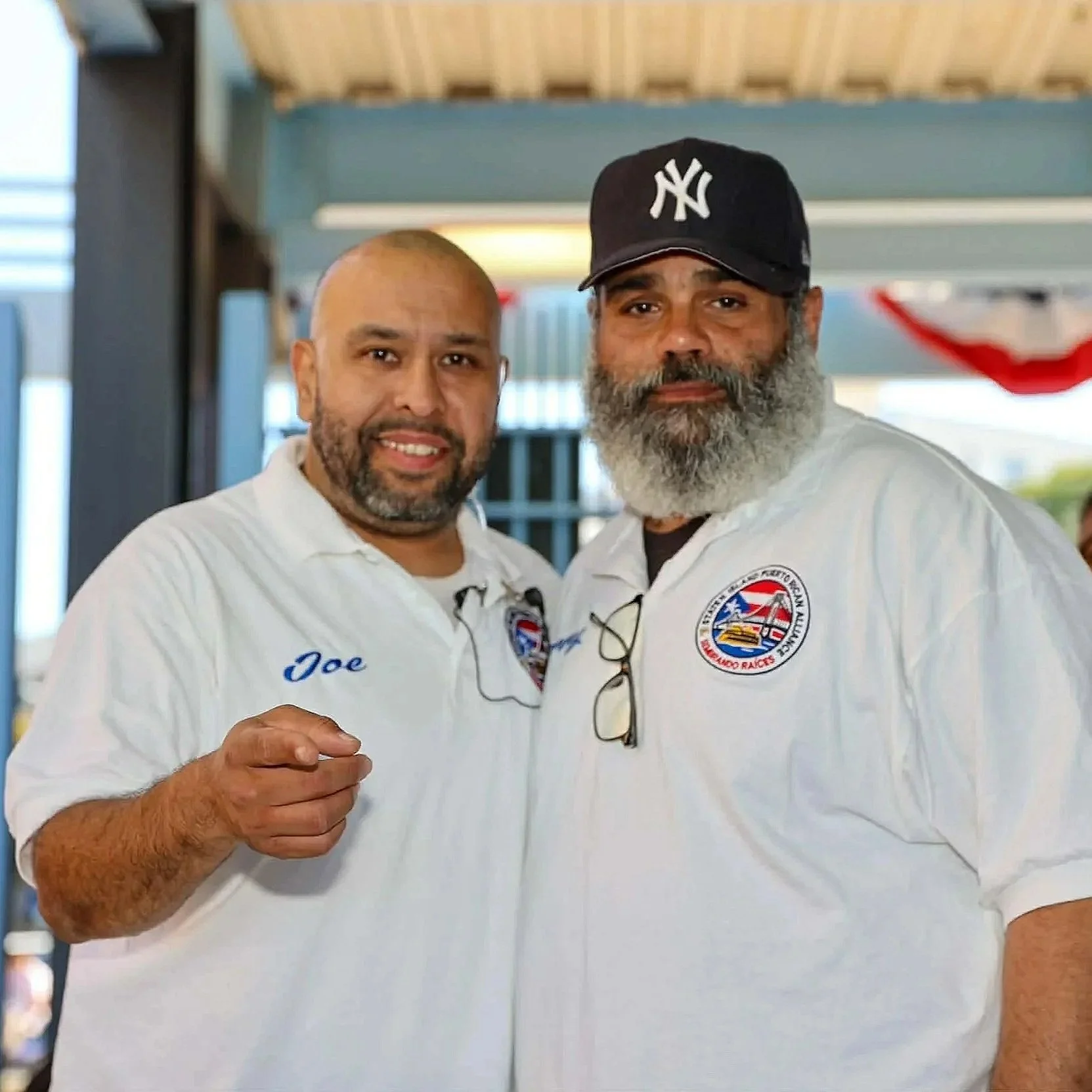 Two men in white shirts with patches pose for a photo together, one wearing a New York Yankees cap. The man on the left has a name tag that reads Joe, a beard, and is smiling while pointing. The man on the right has a full beard, glasses hanging from his shirt collar, and a serious expression.