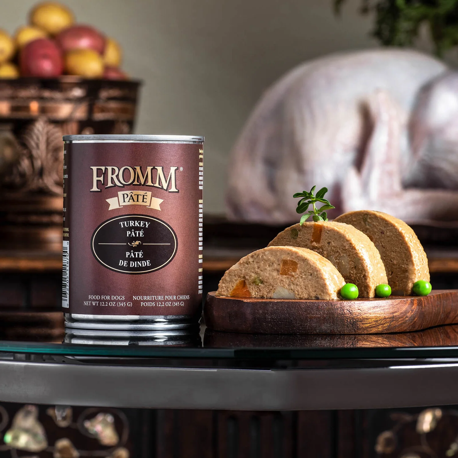 A can of Fromm turkey pâté dog food next to sliced pâté with green peas on a wooden plate, set on a black countertop with woven baskets and a helmet in the background.