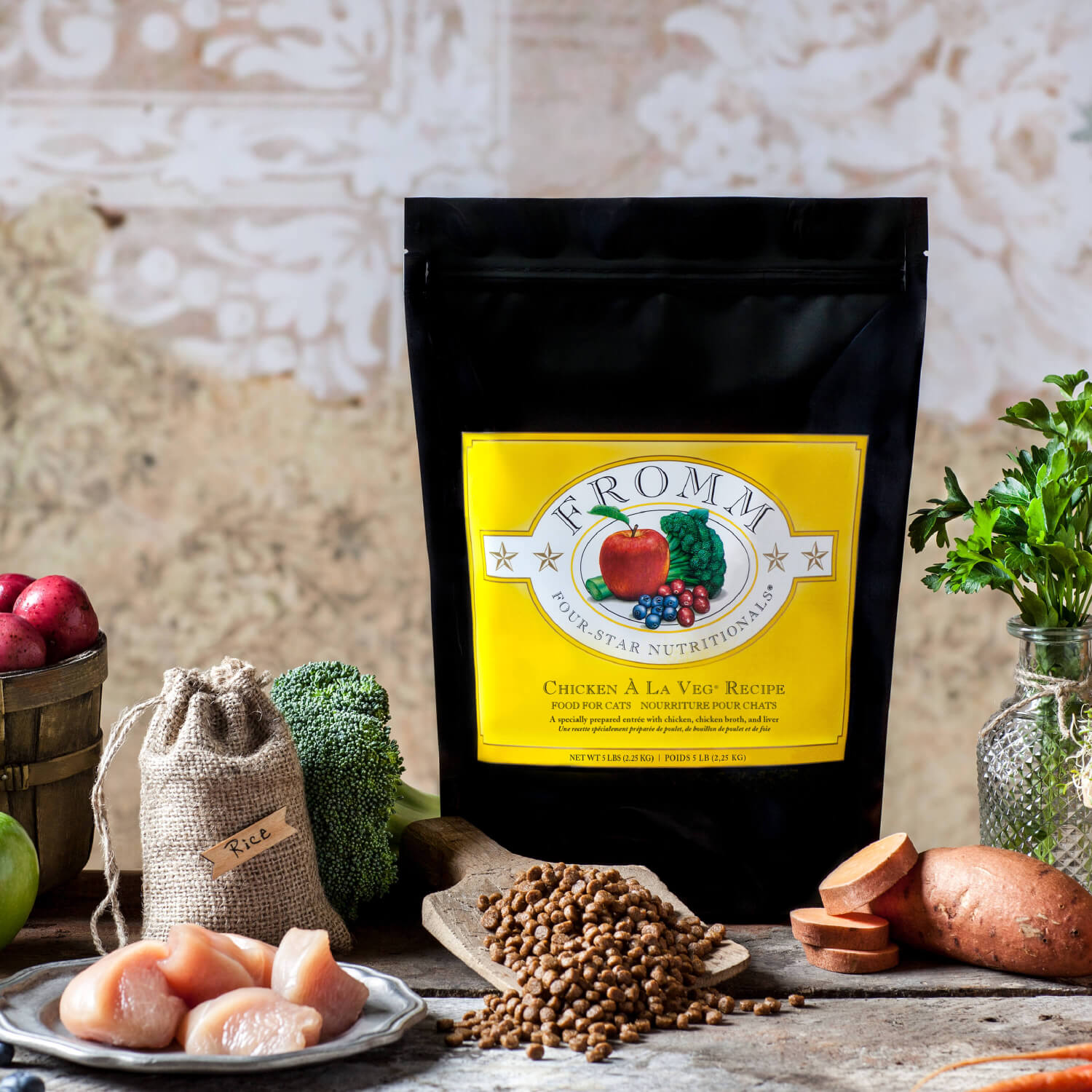 A black package of Faram nutritious cat food labeled 'Chicken A La Veg Recipe' in the center, surrounded by fresh vegetables and ingredients including red potatoes, broccoli, a bag of rice, chicken pieces, sweet potato, and herbs, on a rustic wooden surface with a textured background.
