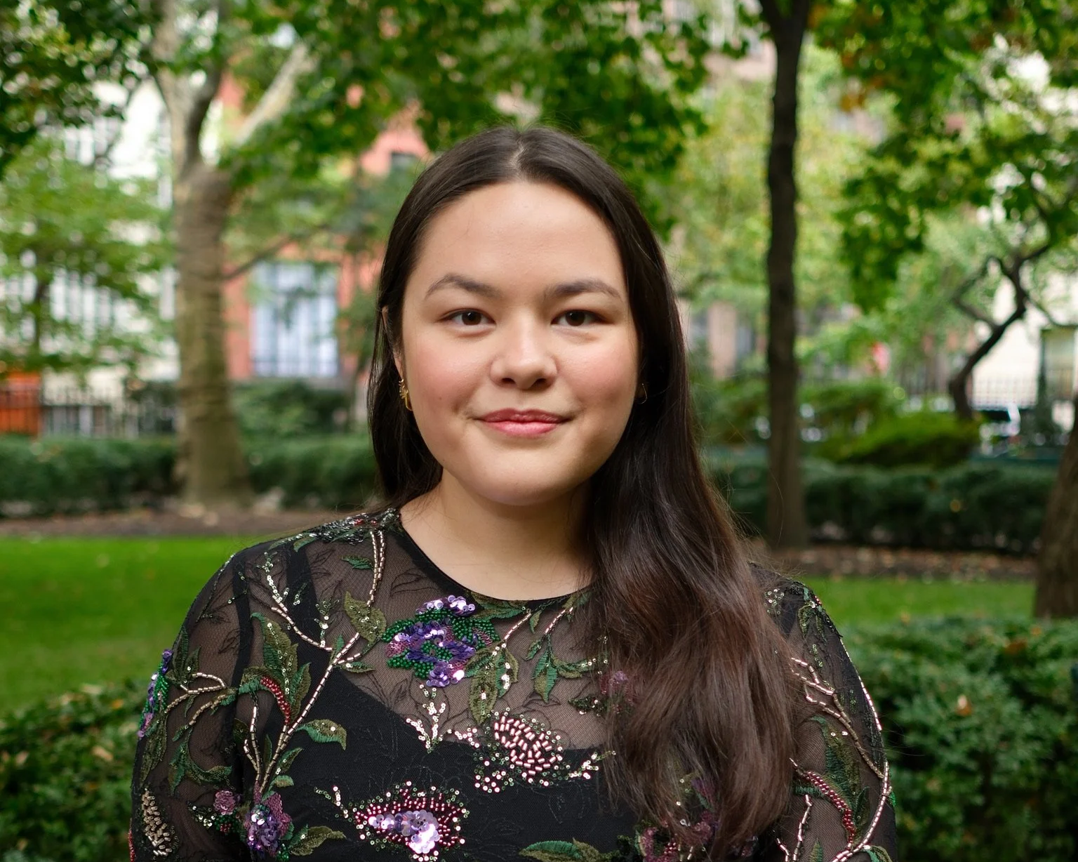 A woman with long dark hair smiling outdoors in a park with trees and buildings in the background.