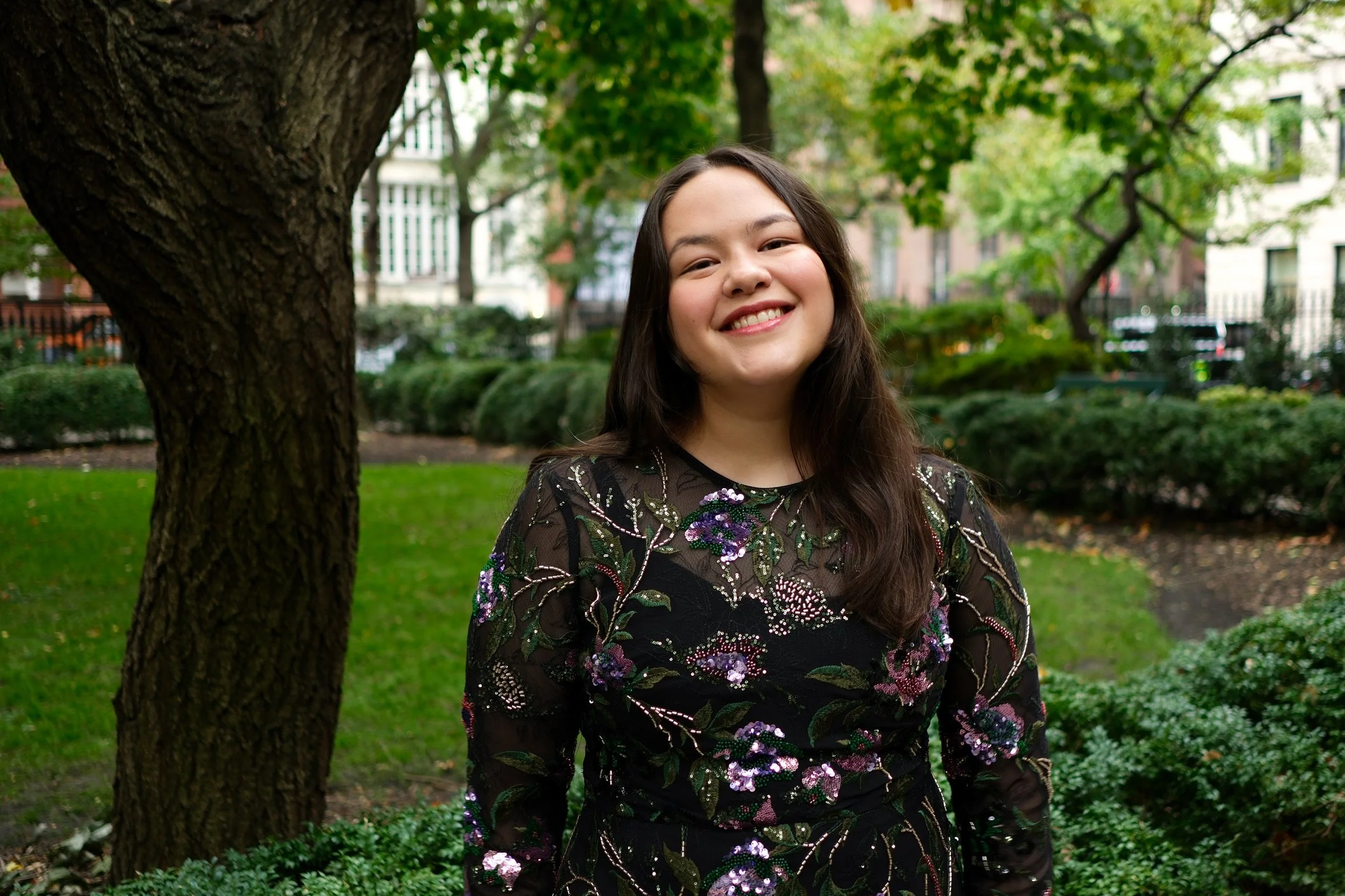 A young woman with long dark hair and a cheerful smile standing outdoors near a tree in a park with green foliage and buildings in the background.