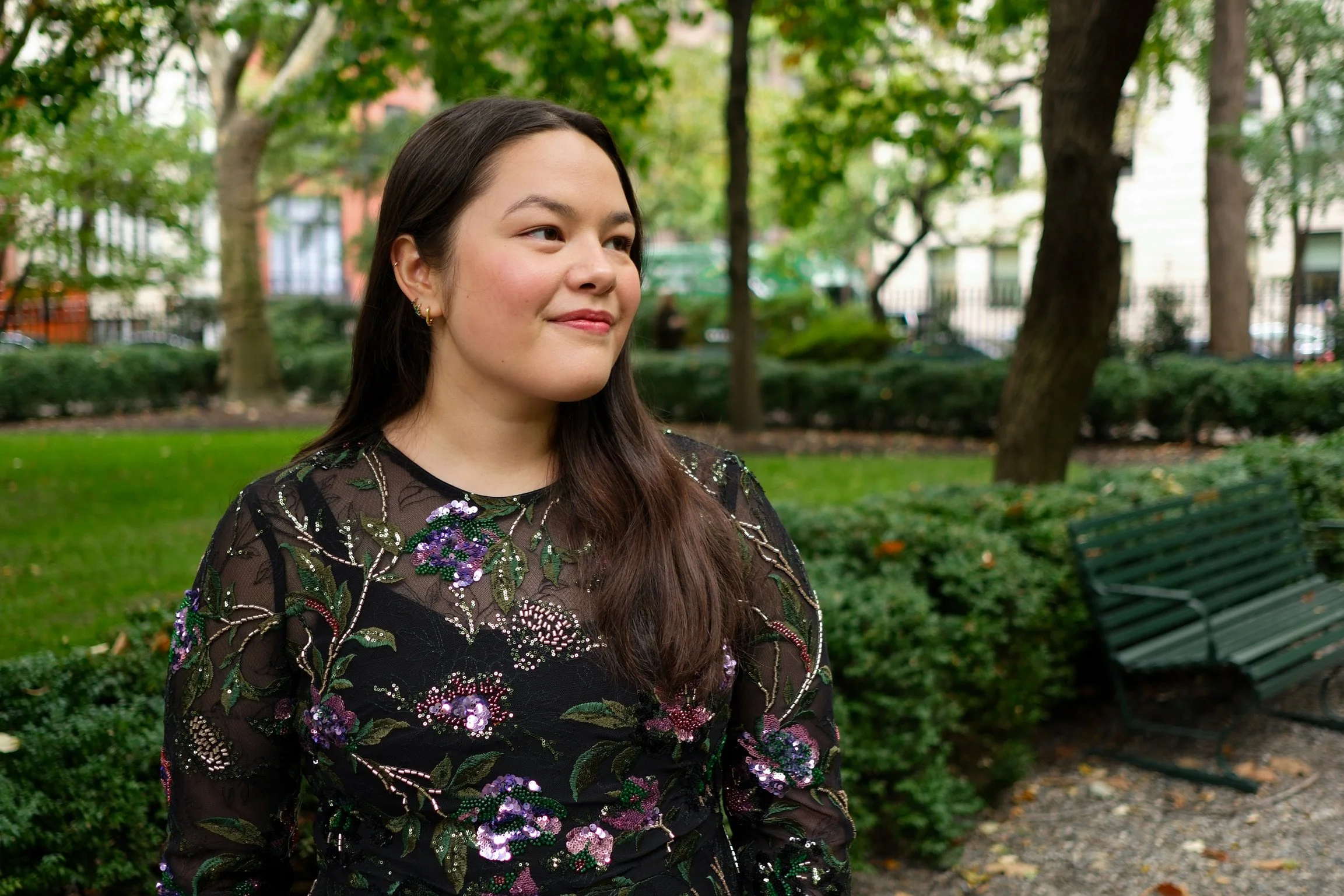 A woman with long dark hair standing in a park with trees and benches, wearing a black dress with colorful floral embroidery.