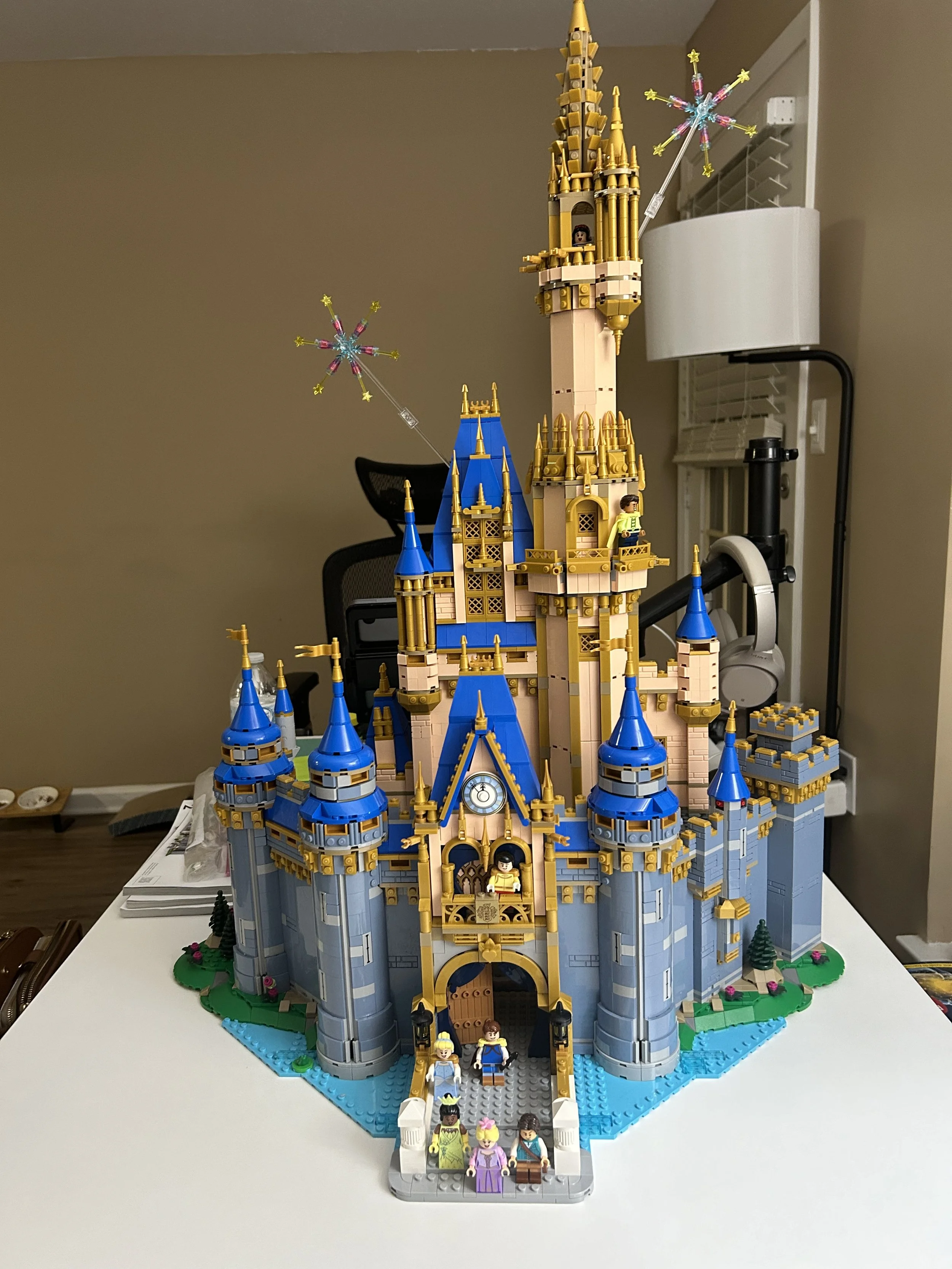 LEGO castle with blue and gray towers, gold accents, and a tall central spire. Several LEGO minifigures are positioned in front and inside the castle, with two floating star-shaped decorations attached to the towers.