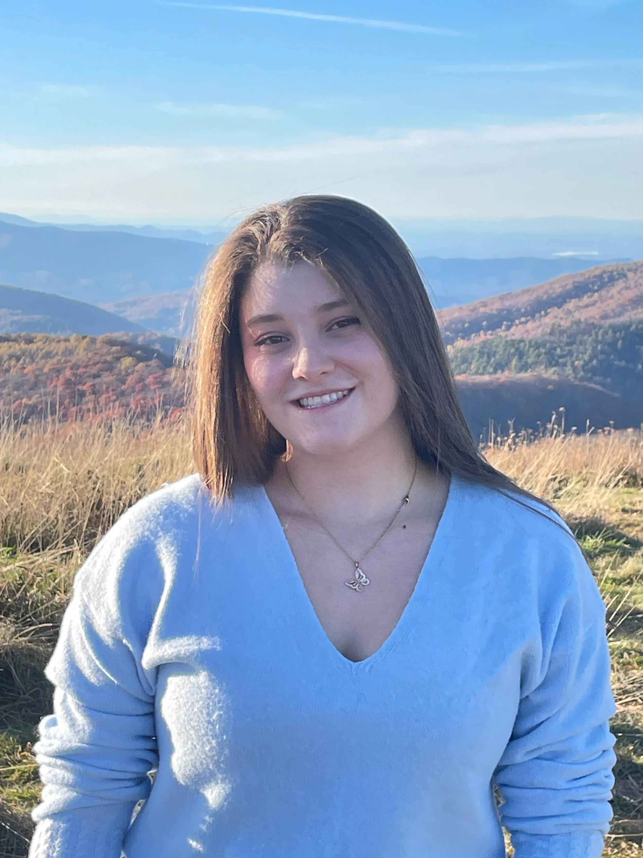 Mariana, a young woman with long brown hair smiling, wearing a light blue sweater and a gold necklace, standing in a grassy hillside with mountains in the background under a blue sky.