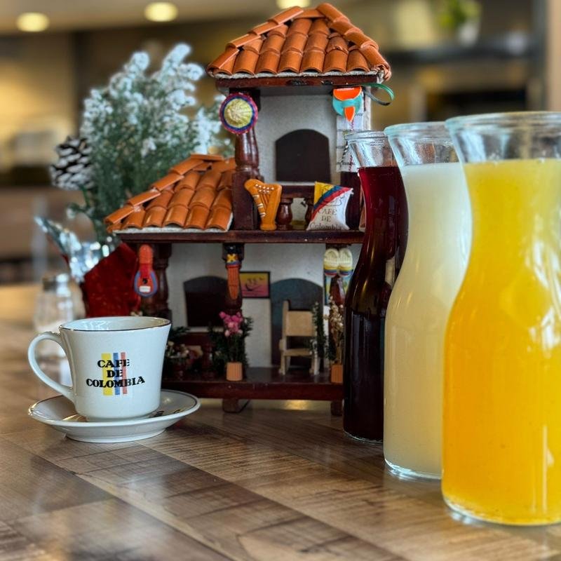 A miniature House on a table with a coffee cup and three glasses of juice in front. The house has a tiled roof, decorated with colorful items, and is surrounded by small plants and furniture traditional from Colombian culture.