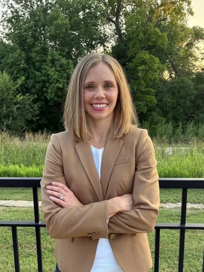 A woman with blonde hair, smiling with arms crossed, stands outdoors in front of a black fence and green trees, wearing a tan blazer over a white top.