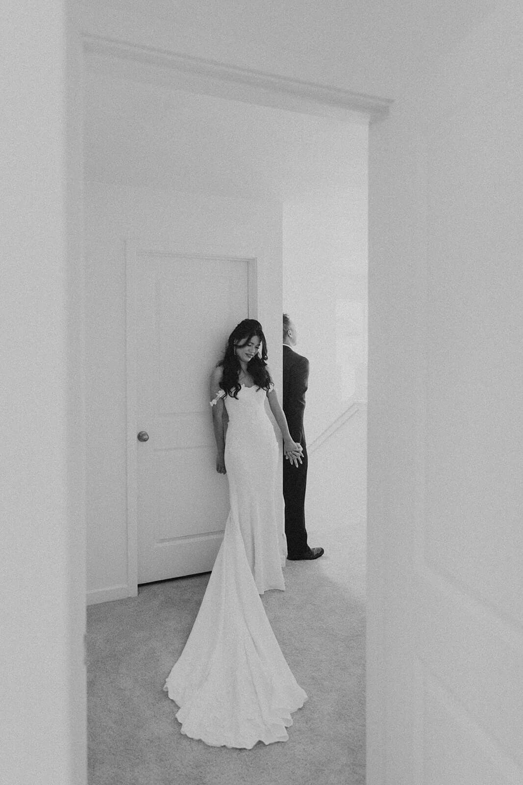 A woman in a wedding dress standing against a wall, holding hands with a man in a suit, who is facing away.