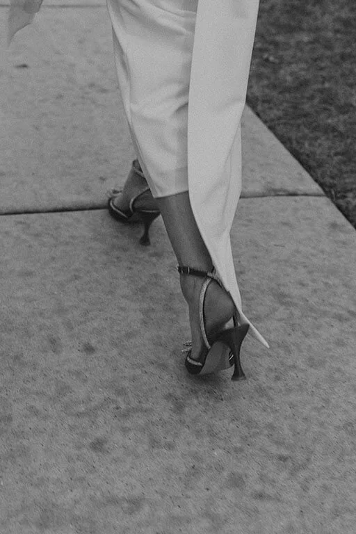 Close-up of a person wearing high heels walking on a sidewalk, with white pants visible.