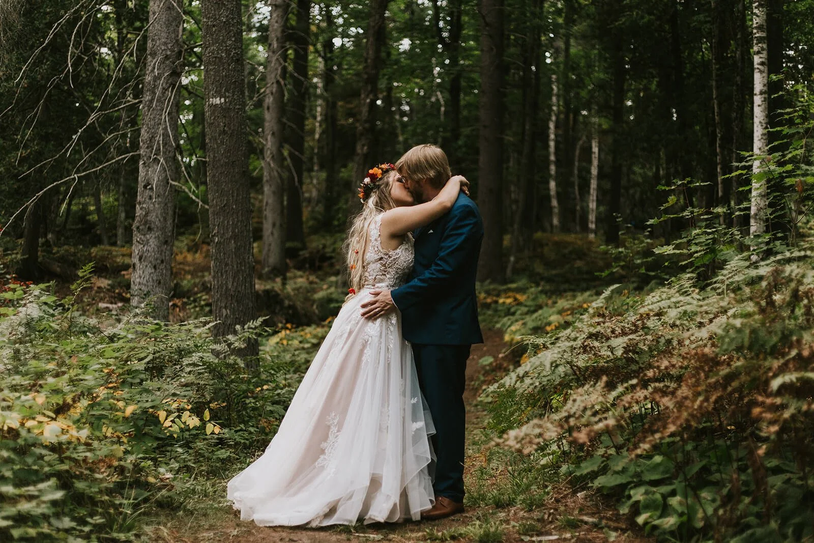 A couple in wedding attire sharing a kiss in a forest with tall trees and green foliage.