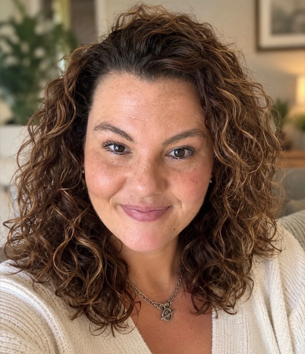 Close-up selfie of a woman with wavy brown hair and light skin, smiling slightly at the camera.