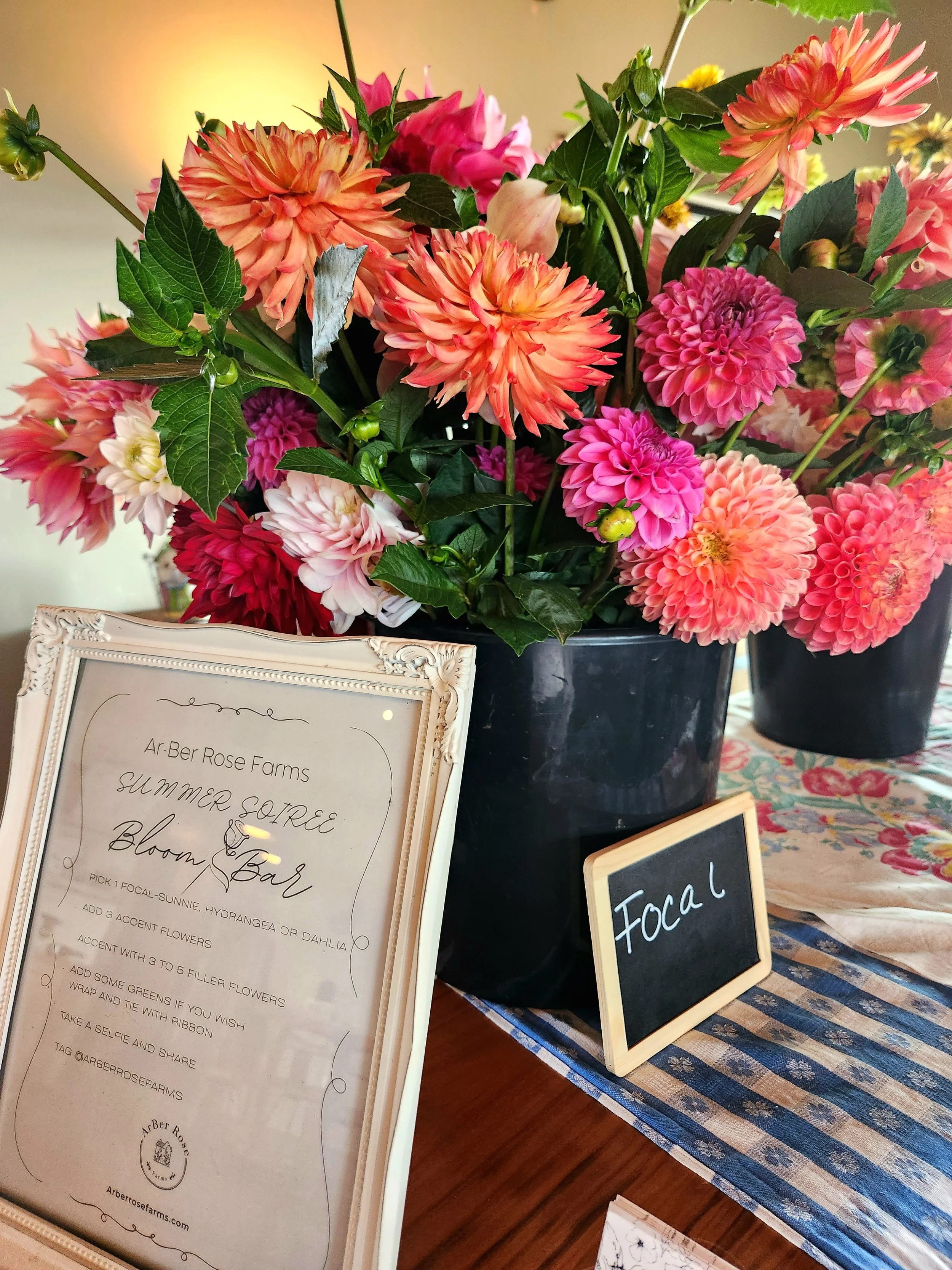 A bouquet of colorful dahlias and other flowers in a black vase, positioned next to a framed sign and a small chalkboard with the word 'Focal' on it, on a table with a patterned tablecloth.