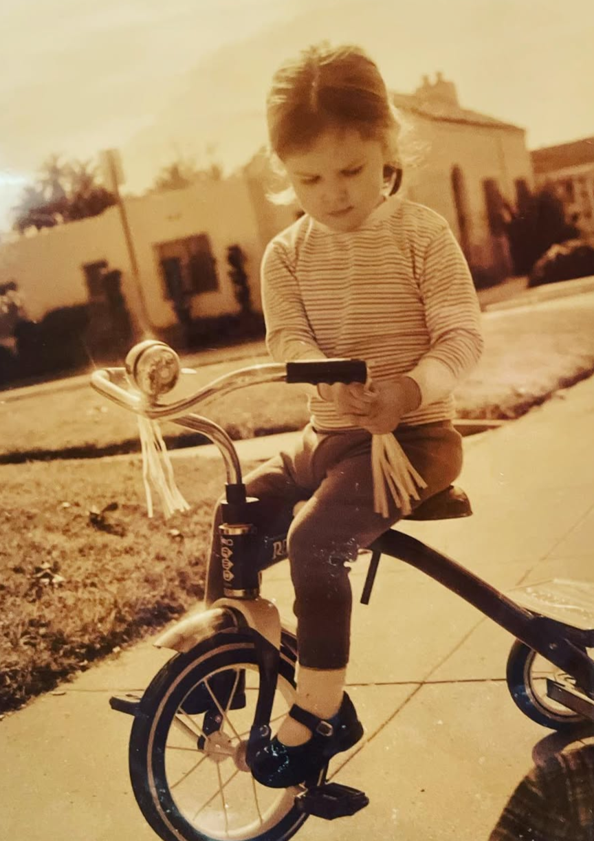 2 year old Eva Luna Ortiz rides a tricycle on Olympic Blvd. Mid-city LA in 2002. The picture is cinematic and sepia.