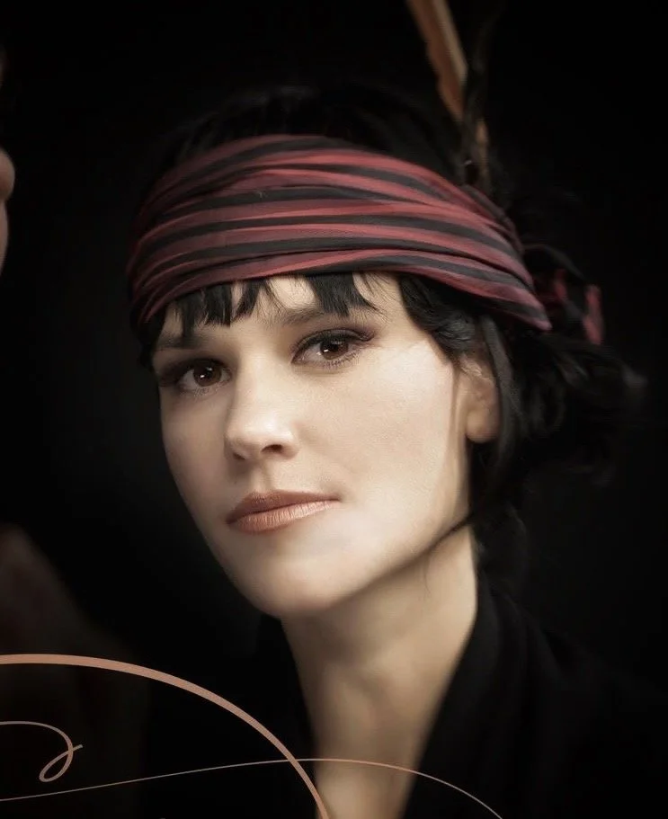Laura Salwet appears in a dramatic cinematic headshot wearing a headband and feather.