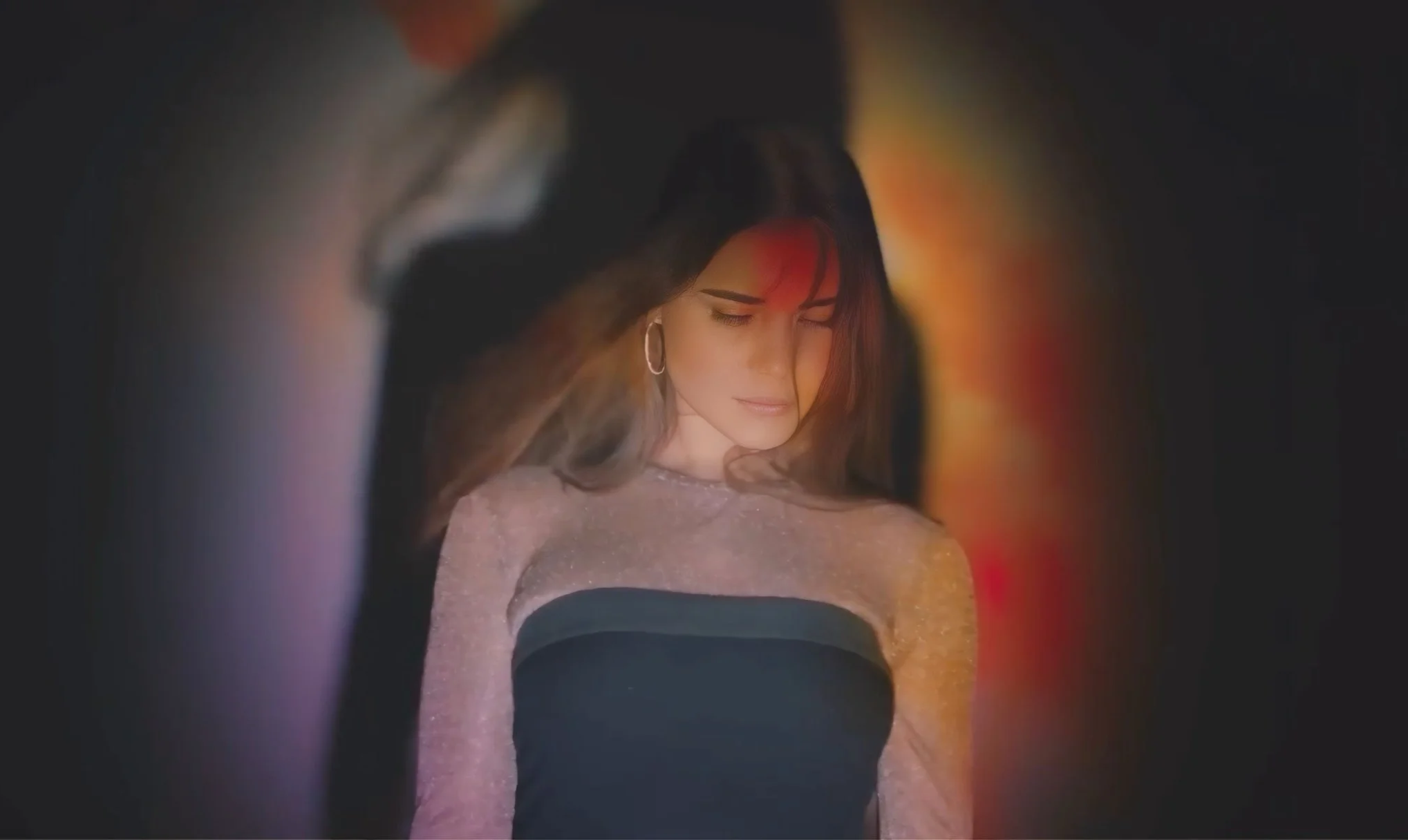 A woman with closed eyes and a content expression, wearing earrings, is seen through a narrow viewing angle with colorful light reflections around her in a dark setting.