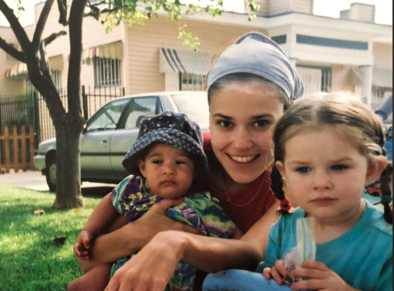 2 year old Eva Luna Ortiz and 1 year old Sarai (now known as influencer and dancer Zoel Esperanza) sit on Laura Salwet's lap in the front yard of her childhood home.
