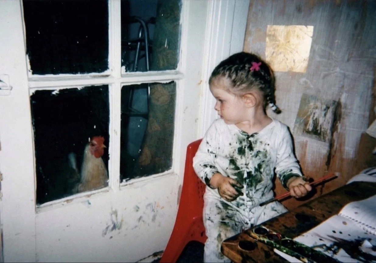 1.5 year old Eva Luna Ortiz is covered in paint while she makes a mess painting with watercolors in a corner desk and stares out a glass door as their backyard pet chicken stares back at her.