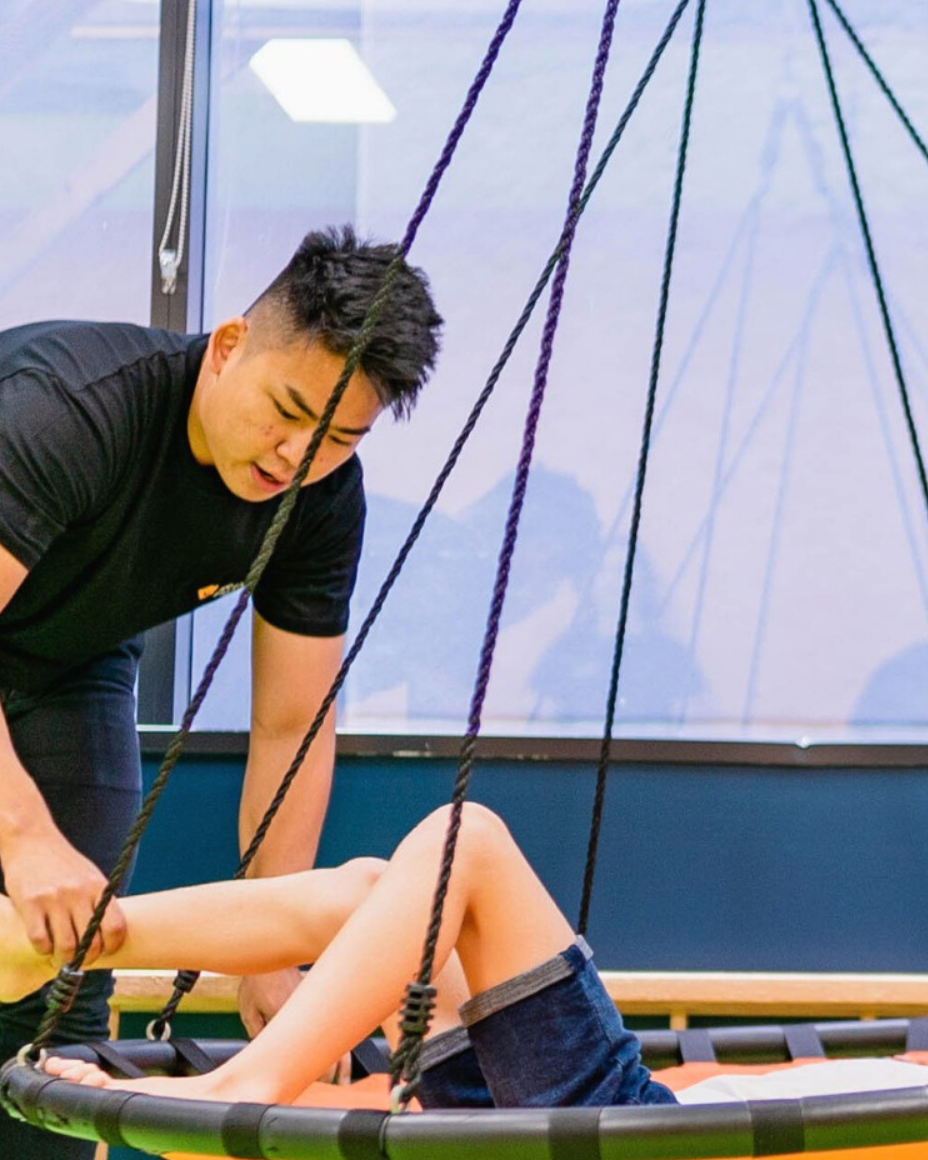 A young man helping a child on a trampoline with a harness and ropes for safety