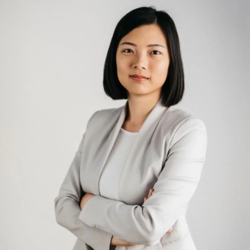 Professional woman with shoulder-length black hair and light makeup, wearing a light gray blazer and white top, standing with arms crossed against a plain gray background.