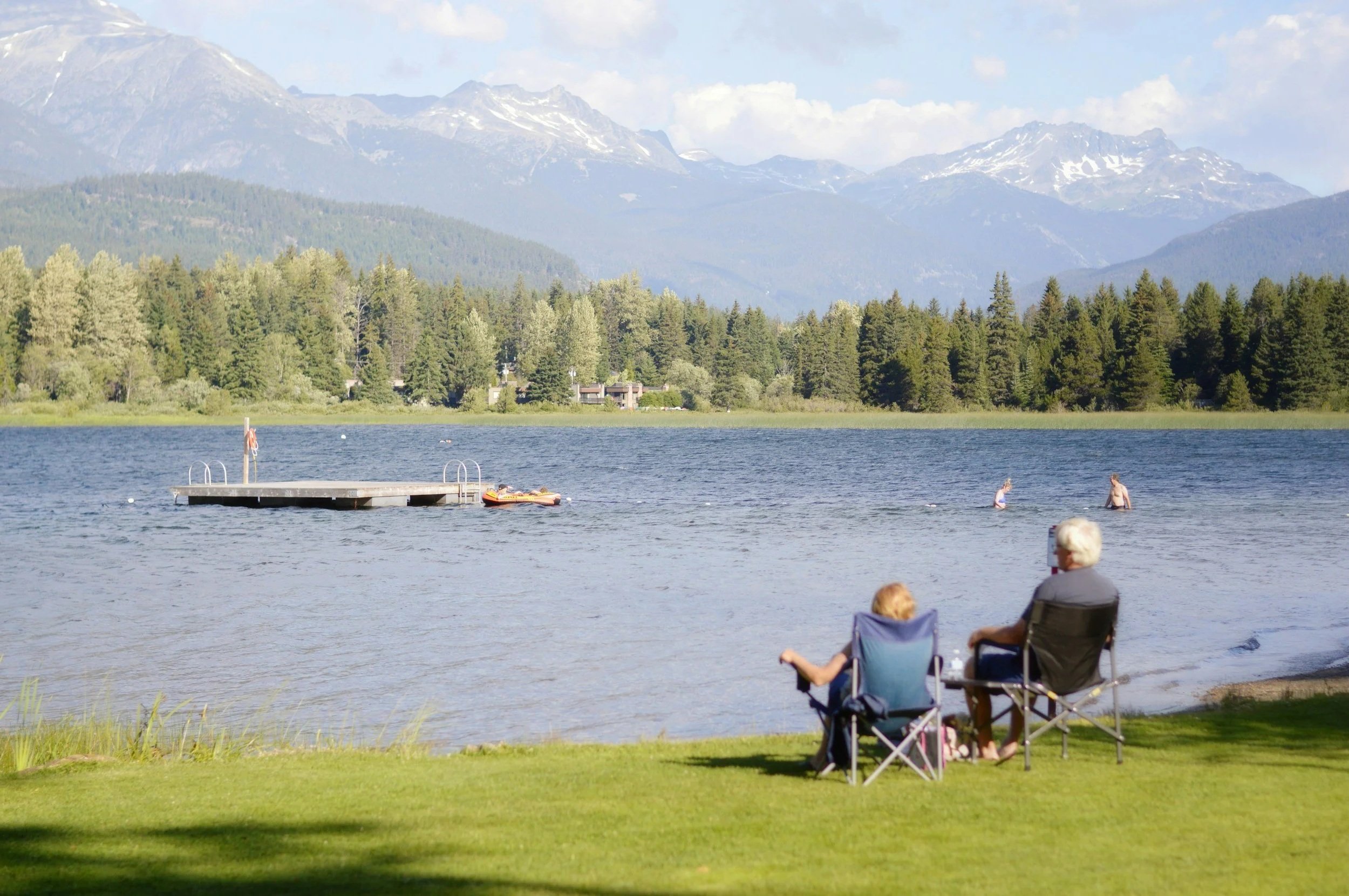 People relaxing on lawn chairs by a lake with a dock, mountains in the background, and trees surrounding the lake.