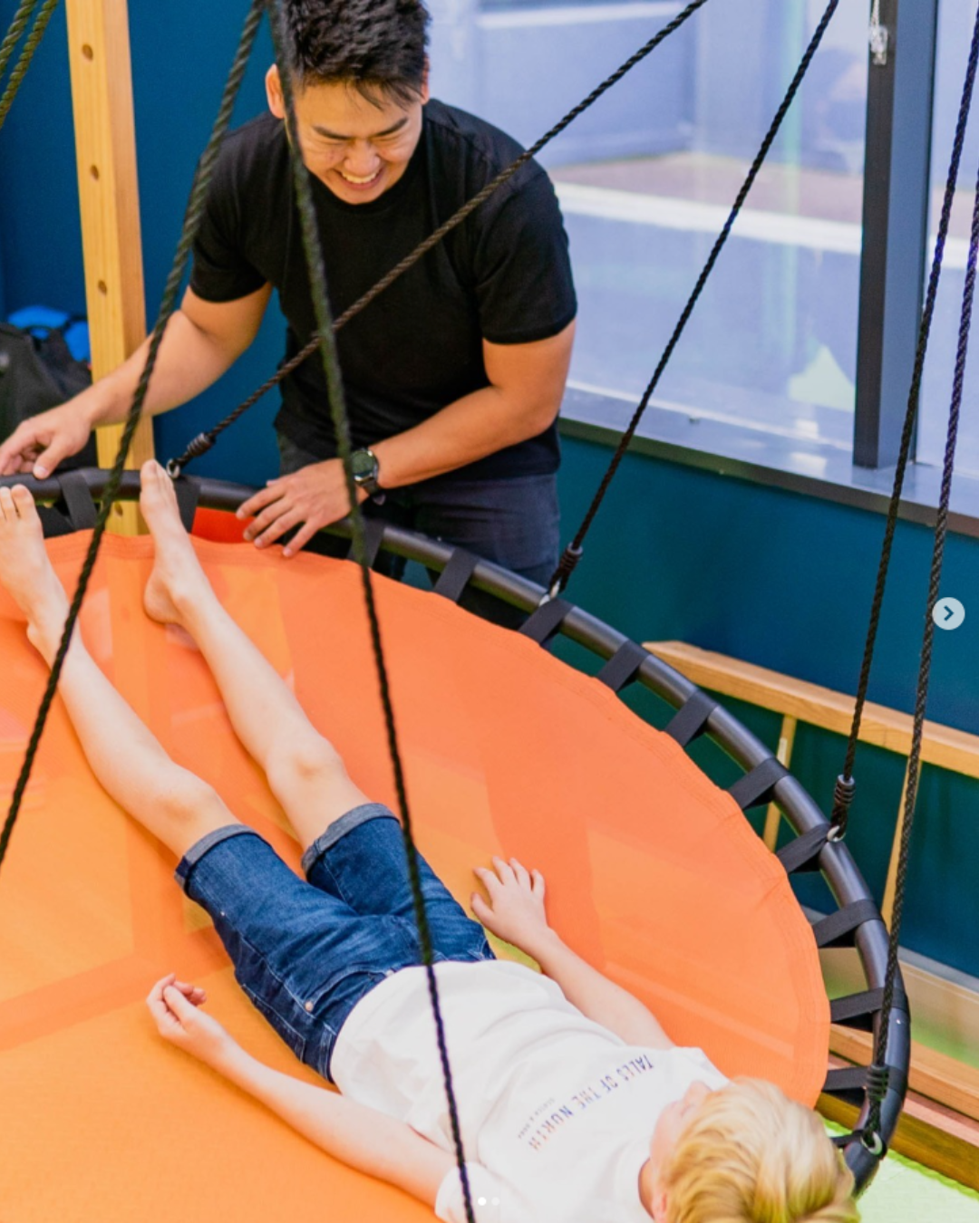 A young boy lying on a trampoline bed with a safety net, while an adult man stands by, smiling.
