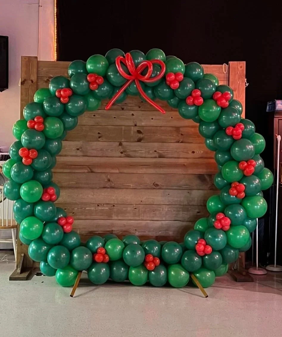 A large holiday arch made of green and red balloons with a wooden backdrop, decorated with a red balloon bow at the top center.