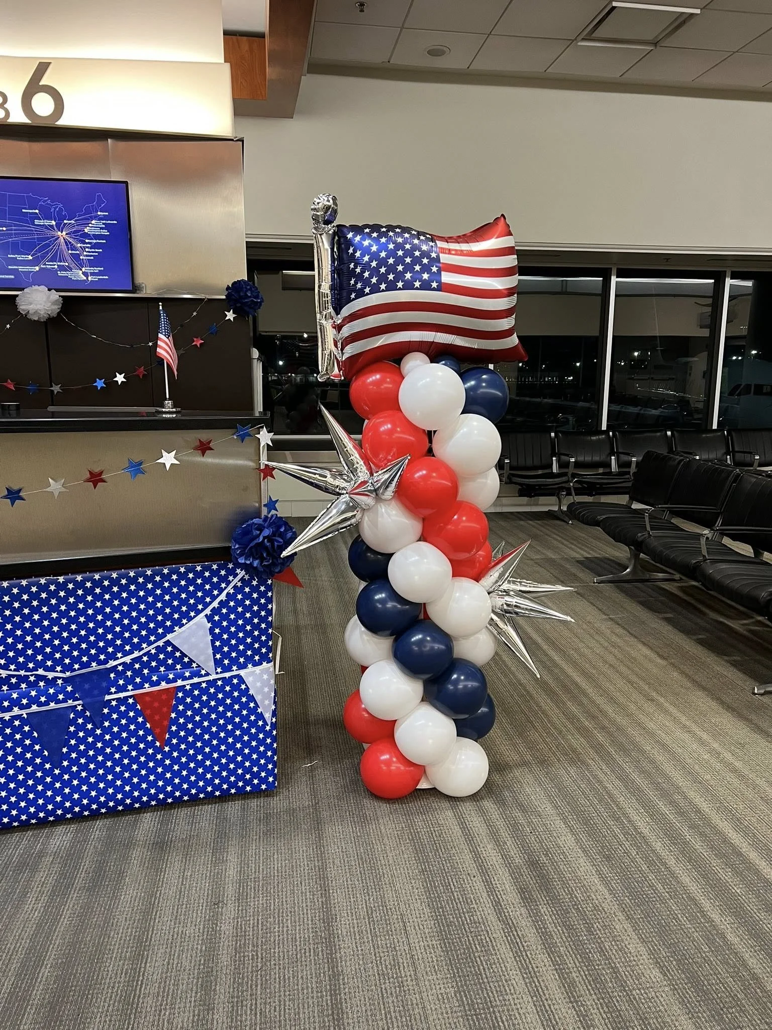 Patriotic decor with a cluster of red, white, and blue balloons, topped with a flag-shaped balloon of the American flag, at an airport or similar setting.