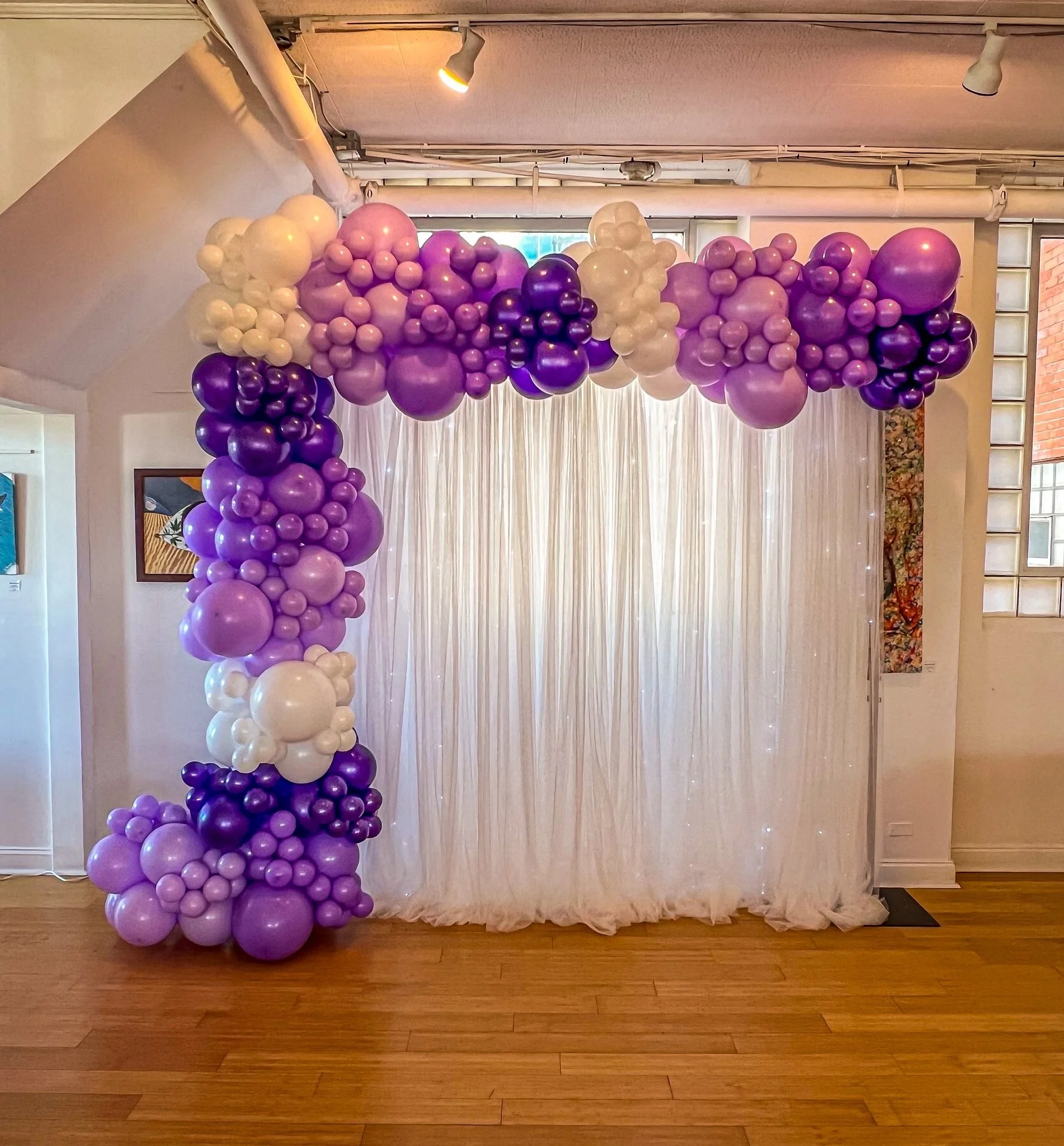 Balloon arch in purple, white, and lavender colors in front of a white curtain backdrop with string lights, inside a room with wooden flooring.