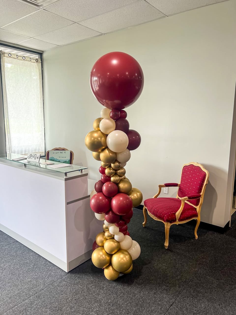 Decorative balloon column with large maroon, gold, and cream-colored balloons, situated near a white reception desk and a red upholstered armchair in an office or event space.