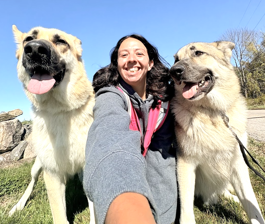 Woman taking a selfie outdoors with two large dogs, both with their tongues out, on a sunny day with clear blue sky and trees in the background.