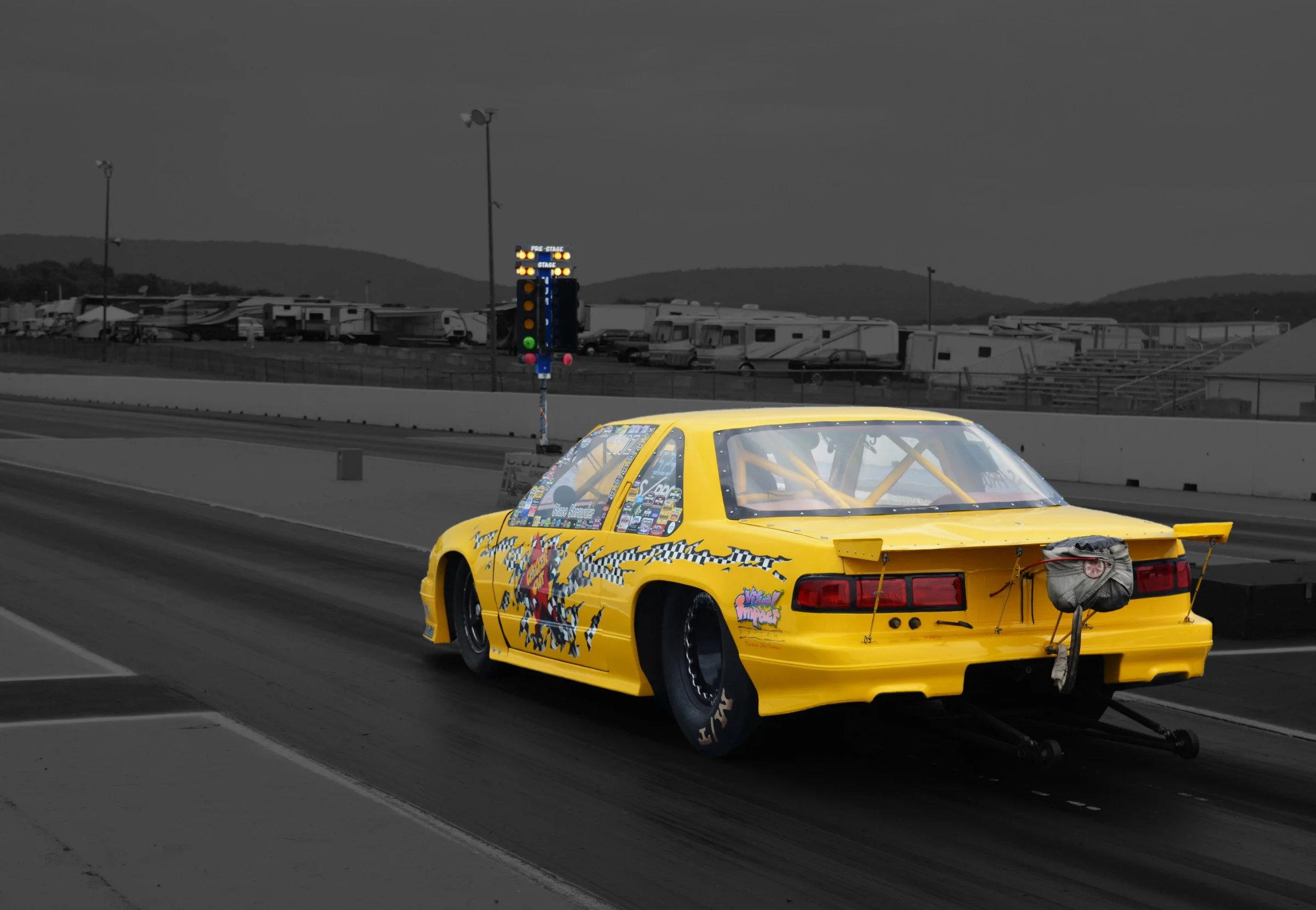 A yellow race car with various stickers parked on a racetrack in front of a drag racing Christmas tree light system. The background shows trailers and RVs with mountains in the distance.