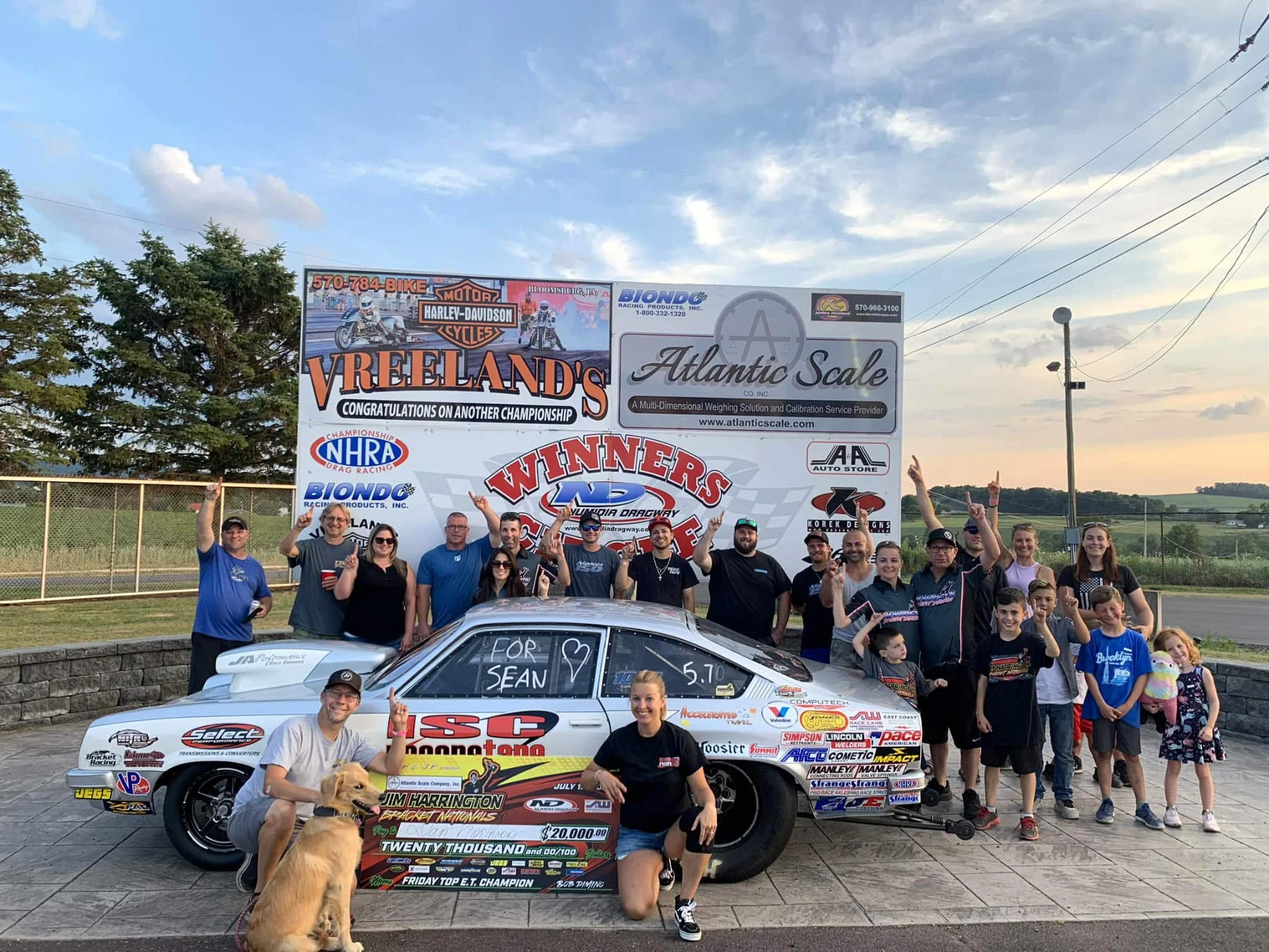 Group of people celebrating at a race track, with a race car in the foreground and a large banner in the background. The race car has various sponsor logos and writing on it, including a large check for twenty thousand dollars awarded to Jim Harringt
