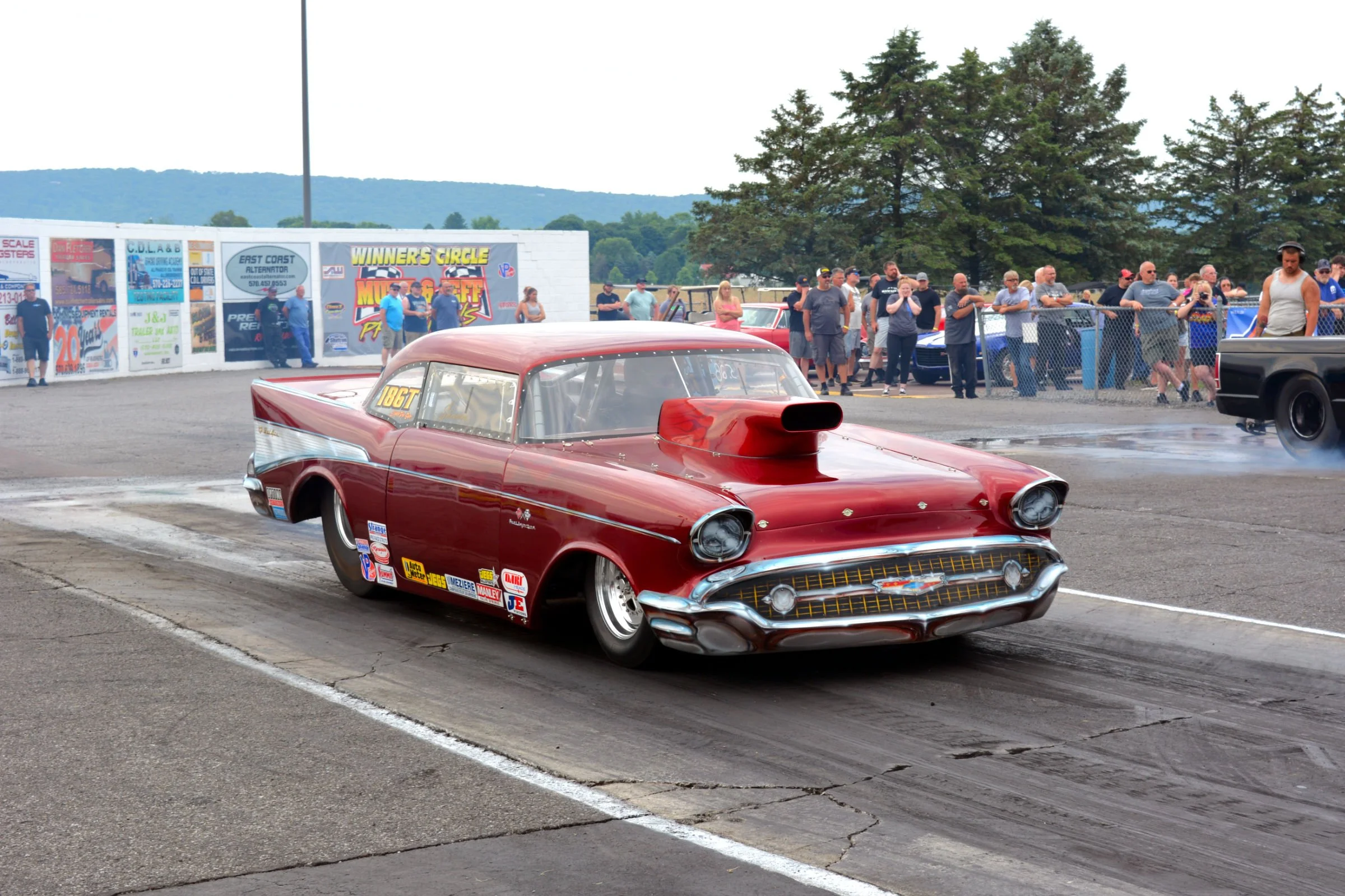 A red classic drag racing car with a large hood scoop on the starting line at a drag strip, with a crowd of spectators watching behind a safety fence, and trees and hills in the background.