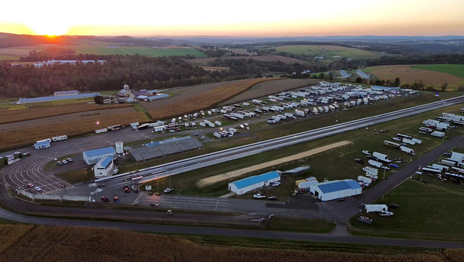 A large outdoor racetrack with parking lots full of RVs and vehicles, surrounded by farmland at sunset.