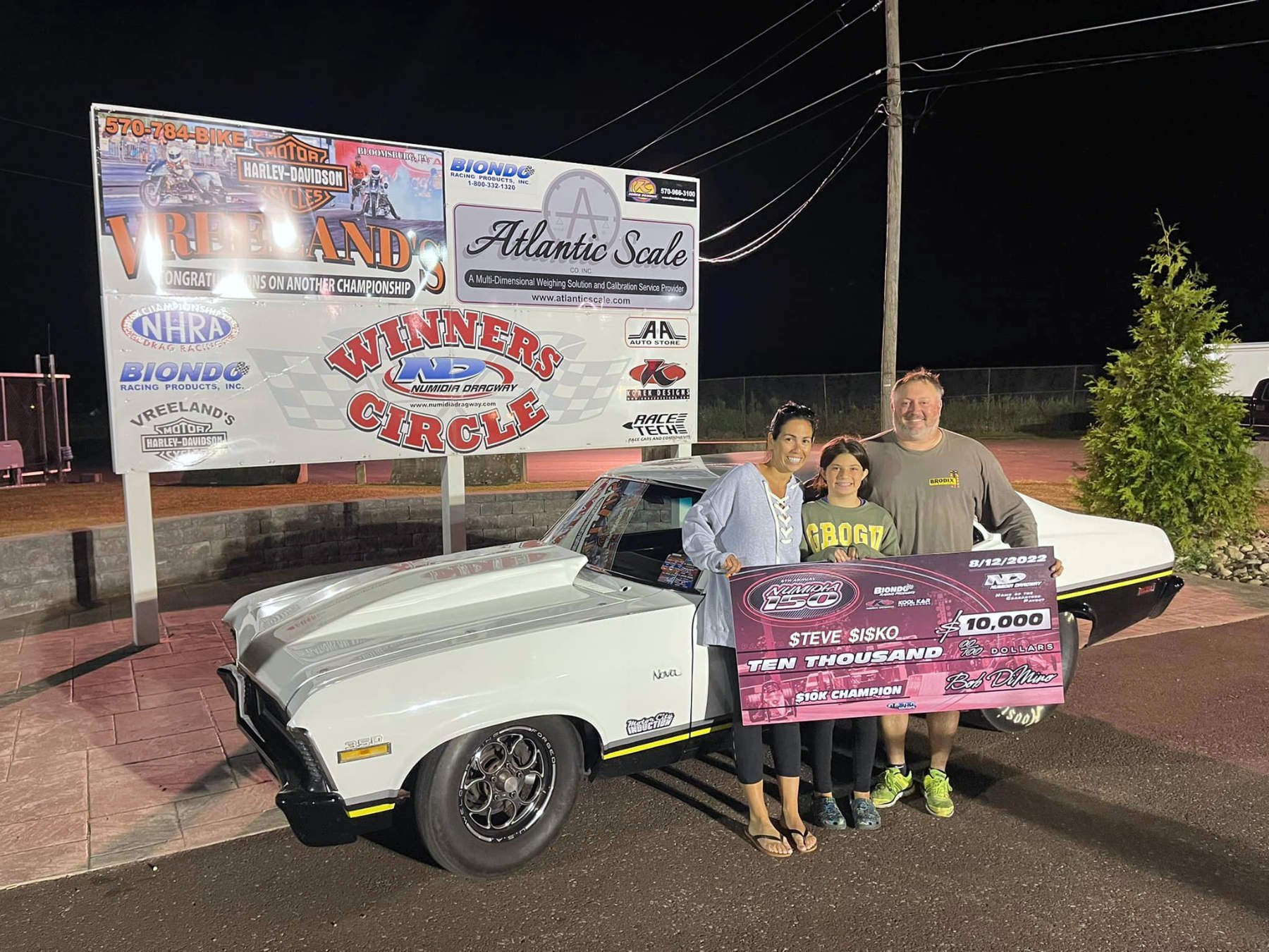 Three people standing next to a white car with a large check for ten thousand dollars and a trophy in front of a promotional banner for a drag racing event. The banner displays various sponsors and a motorcycle race image.