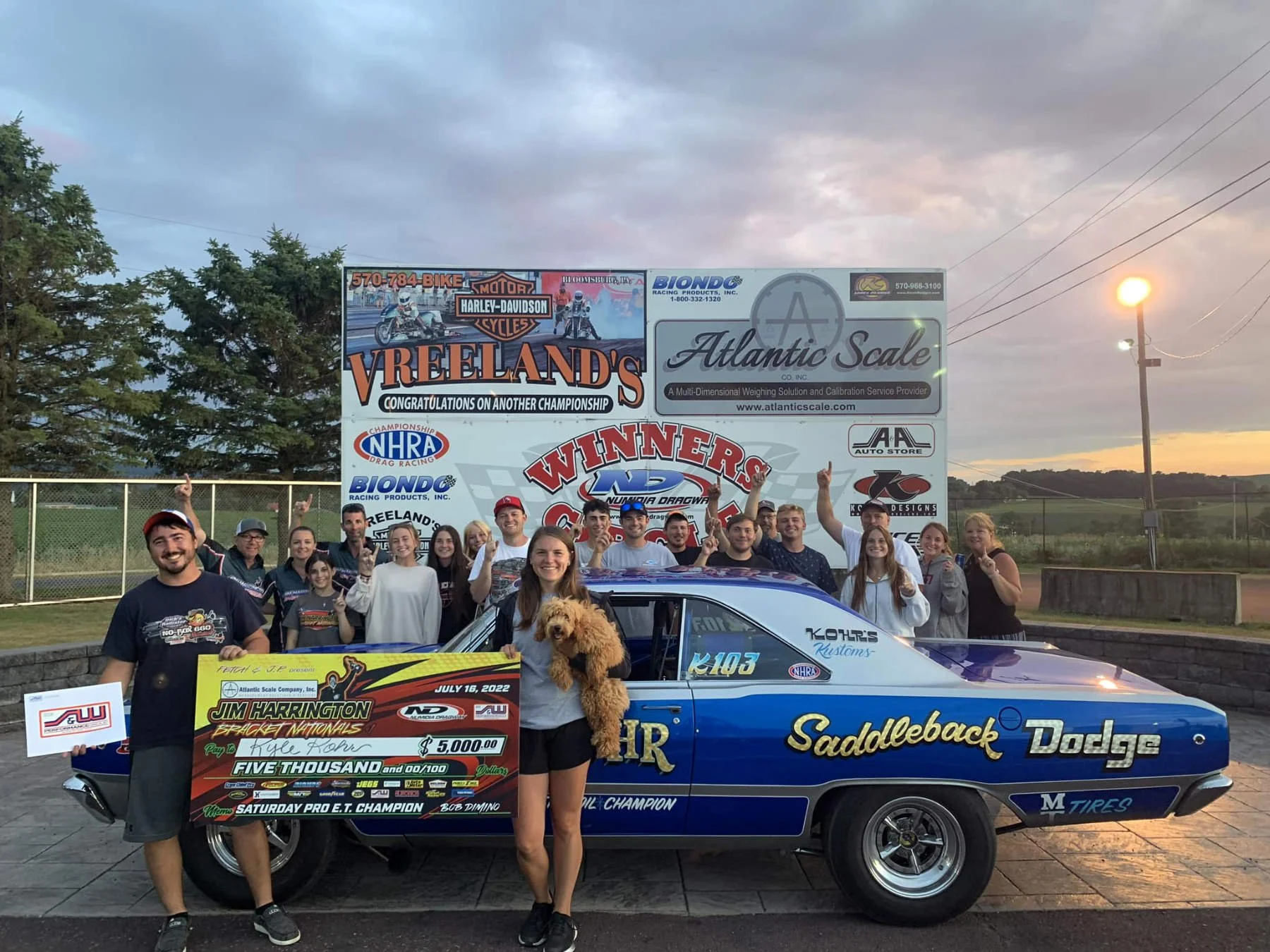 Group of people celebrating at a racing event, standing in front of a blue Dodge car with sponsorship decals. One person is holding a large check for $5,000 awarded for winning a drag racing event, and a woman is holding a small dog. The background f