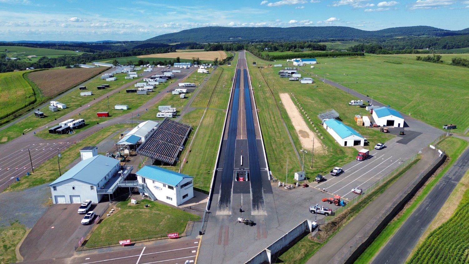 Aerial view of a rural drag strip with racing lanes, surrounded by farm buildings, trailers, and open fields, with hills in the background.