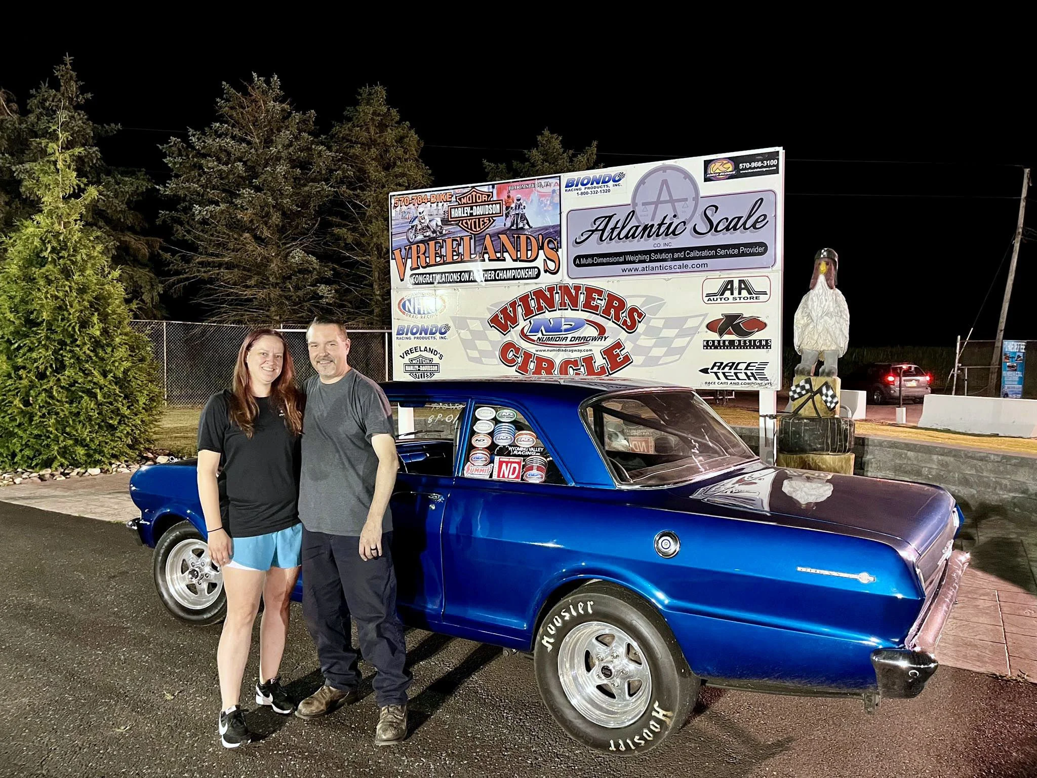A man and woman standing next to a blue car at night with advertising signs and trees in the background.
