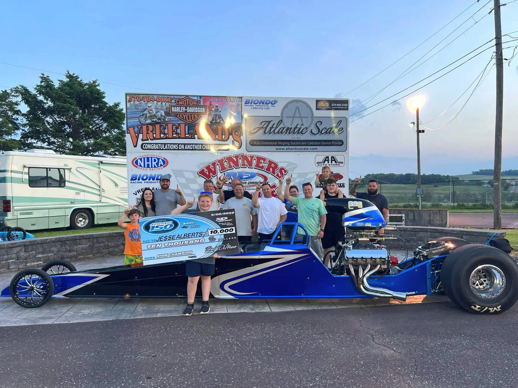 Group of people celebrating with a drag racing car and a large check for $10,000 at a raceway, with banners and a trailer in the background.