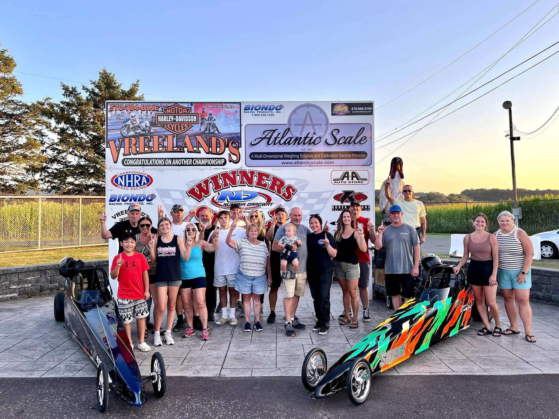 Group of people outdoors at a racing event, posing in front of a large advertising billboard, with race cars and racing paraphernalia in the foreground, and a racing track and open sky in the background.