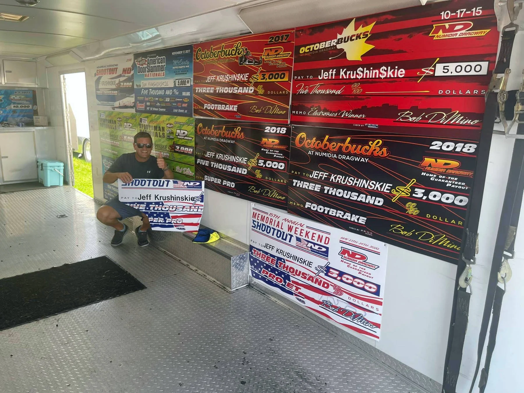 A man kneeling inside a trailer holding a large replica check. The trailer's walls are decorated with multiple large banners and posters related to drag racing and awards, featuring names like Jeff Krushinske, Bob DiMino, and the event Octoberbucks a