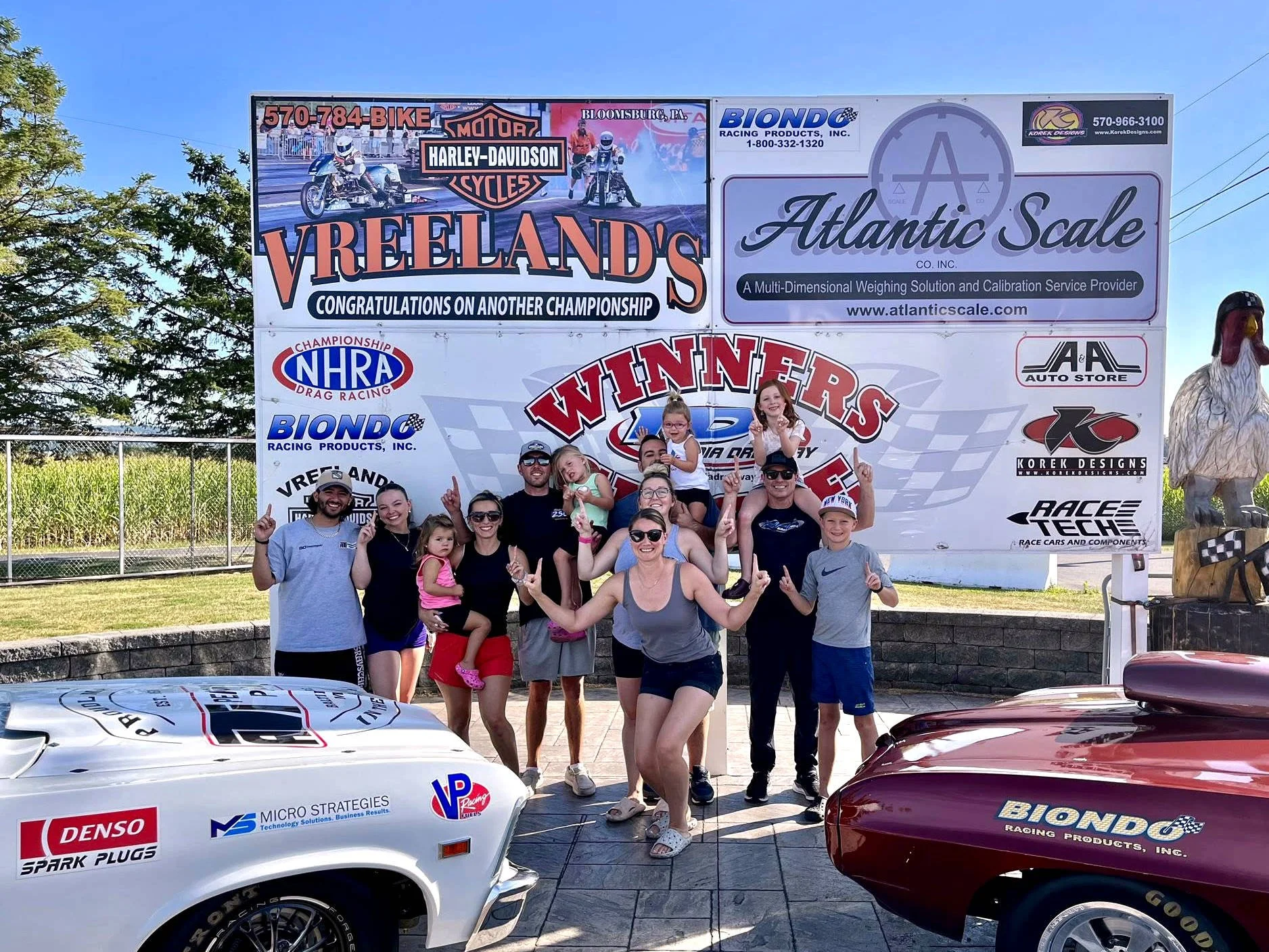 A group of people, including children and adults, gathered in front of racing cars and large banners at a drag racing event. The banners display logos and sponsors, indicating a championships event at Vreeland's HD Harley-Davidson and Atlantic Scale.