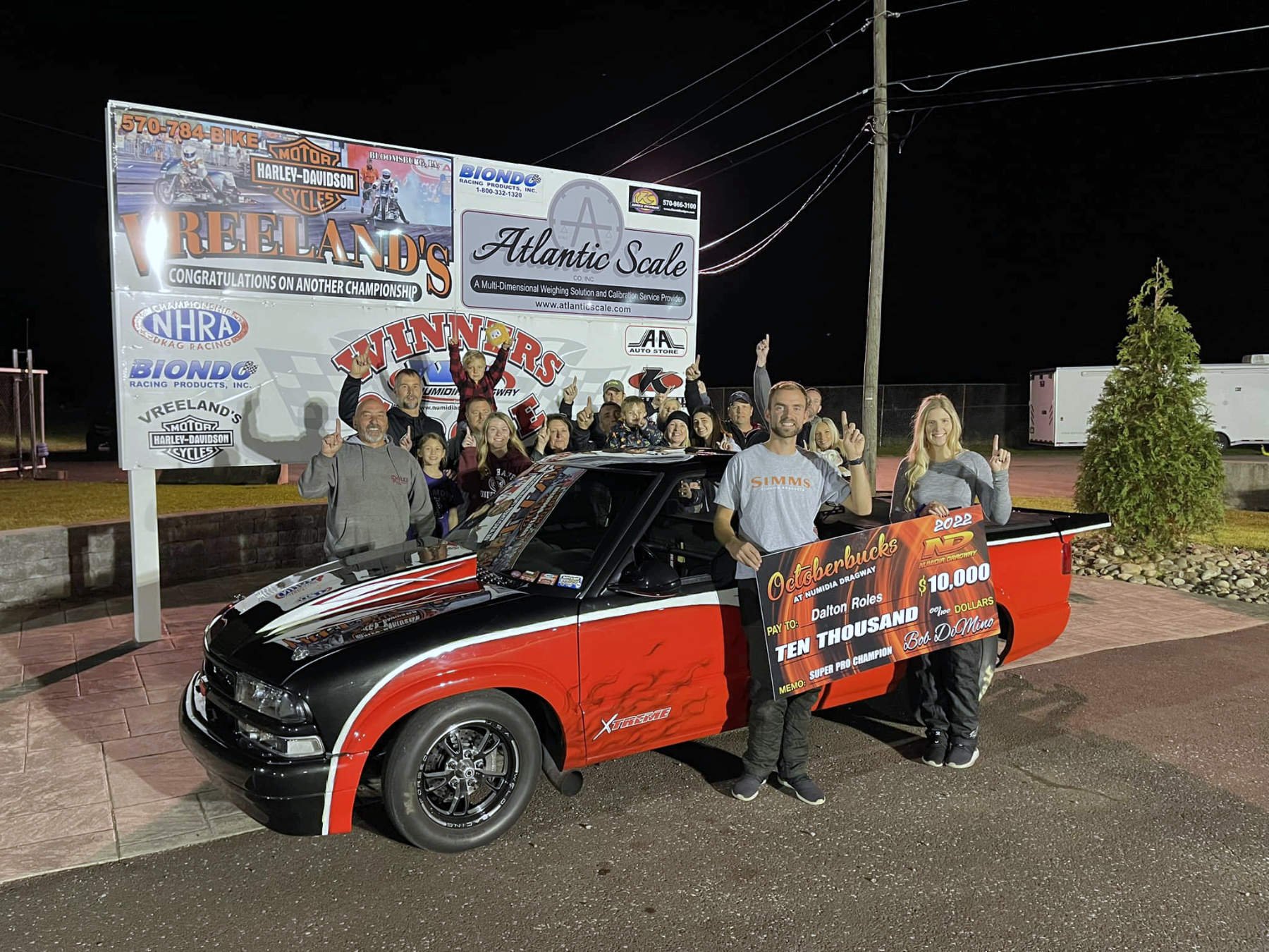 Group of people celebrating outdoors at night around a red and black race car, holding a large check for ten thousand dollars, with a sign displaying various sponsors and a victory banner in the background.