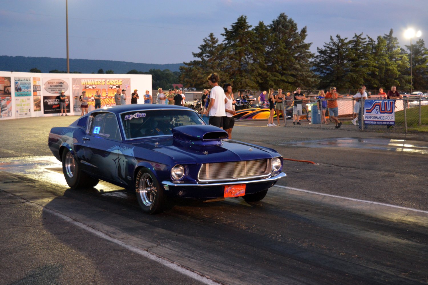 A blue drag racing car with a hood scoop at a racetrack during evening, with people standing and watching in the background.